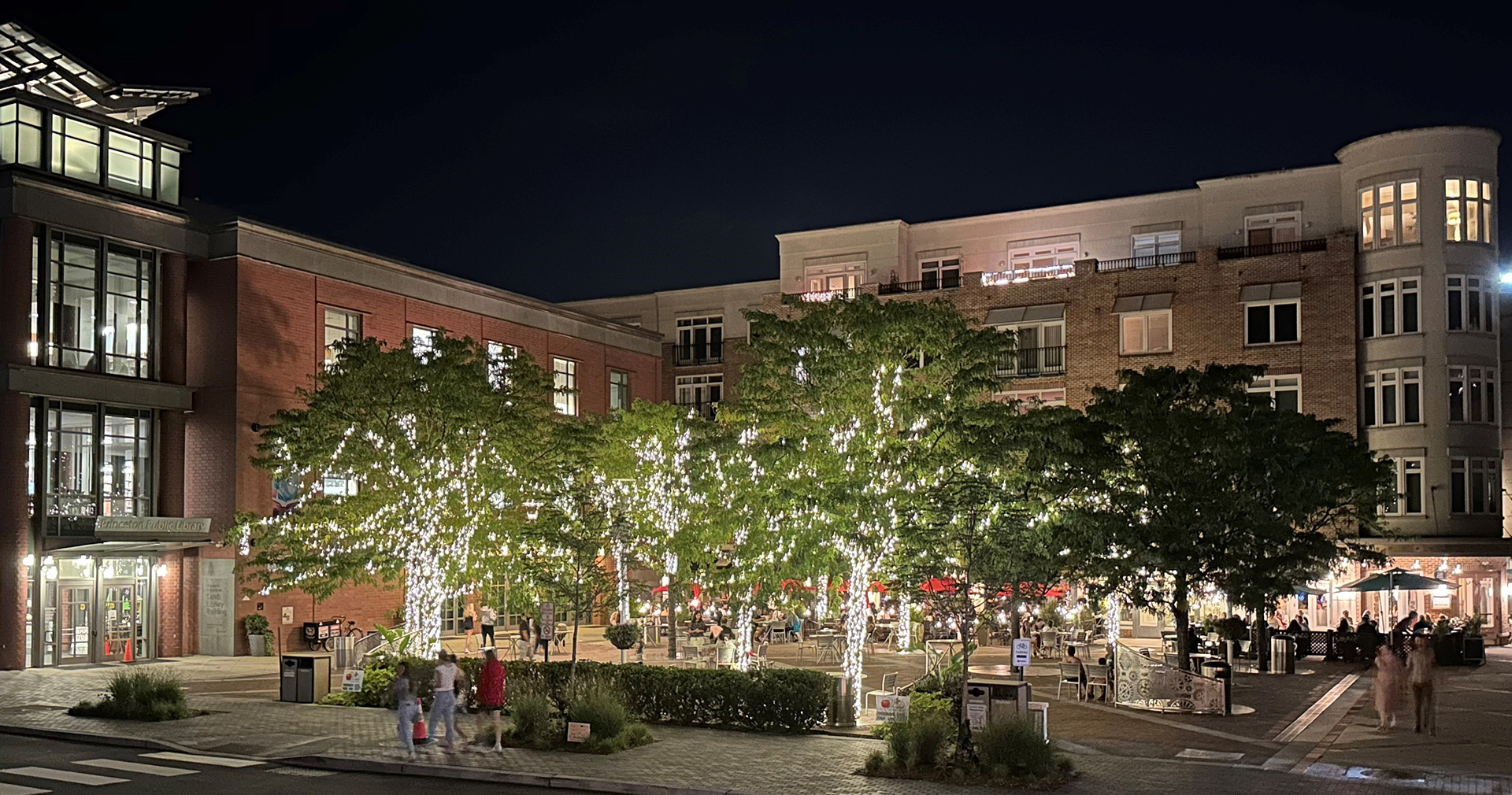 Nighttime view of urban courtyard space surrounded by buildings with several mature trees lit up with lights and people walking by.